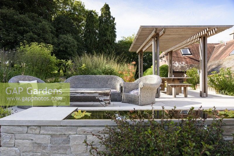 Terrasse dans un jardin de campagne en juillet avec meubles en osier et belvédère en bois vu au-delà d'un plan d'eau 