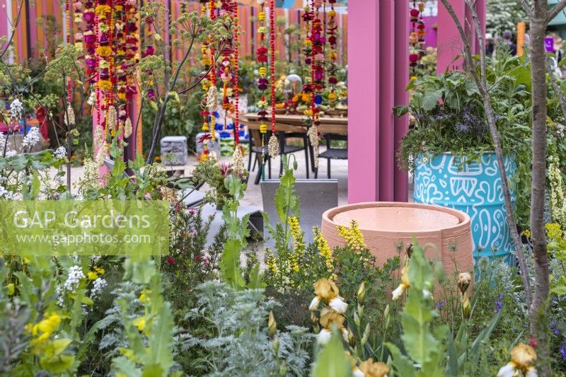Une vue à travers un parterre de fleurs vivaces jusqu'à un plan d'eau circulaire rose pastel à côté d'un baril de pétrole peint en bleu et blanc planté de fleurs et de légumes dans le jardin d'unité du RHS et de l'Eastern Eye. Créateur : Manoj Malde 