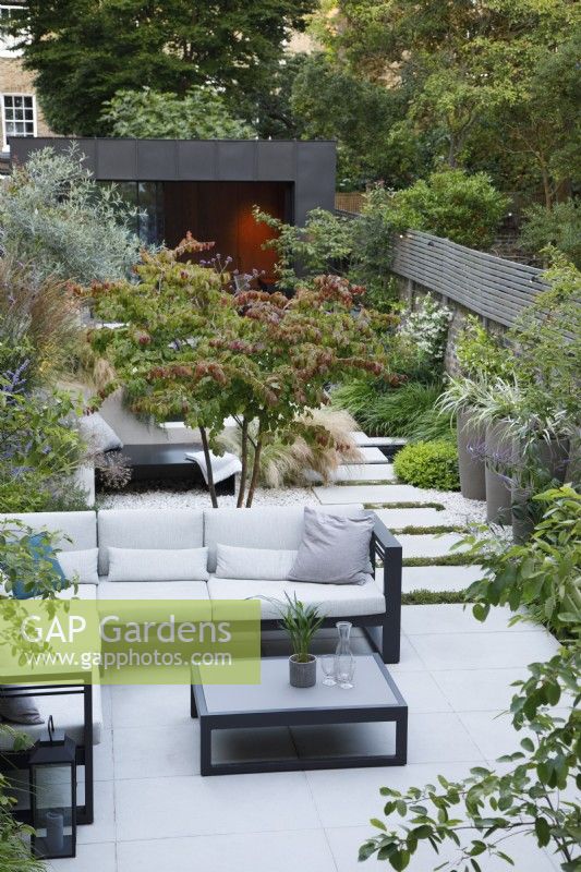 Vue d'ensemble de ce jardin de ville avec le coin salon avec Parrotia persica multi-troncs, chaise longue et chemin menant au bâtiment recouvert de zinc 