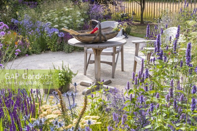 Coin salon avec table en bois et deux chaises entourées de plantes vivaces à floraison estivale dont Salvia nemerosa 'Caradonna', Agastache 'Black Adder' et Eryngium. Jardin des héros horticoles emblématiques RHS, concepteur : Carol Klein, RHS Hampton Court Palace Garden Festival 2023 