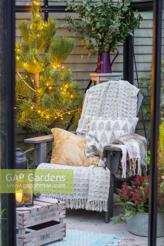 Chaise en plastique recyclé avec couvertures et coussins, caisse en bois et plantes ornées de guirlandes lumineuses à l'intérieur d'une serre 