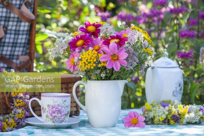 Bouquet d'été contenant du Dahlia, du Tanacetum vulgare et de l'Astrantia dans un vase blanc, une couronne de fleurs, une valise pique-nique et un service à thé. 