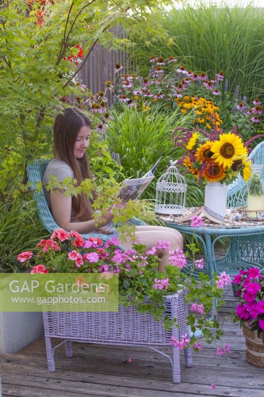 Une jeune femme aime lire un magazine sur une terrasse d’été ornée de pots plantés de pélargoniums. 