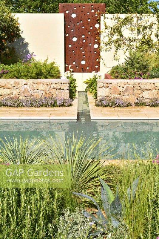 Mur avec panneau en acier avec une cascade d'eau qui se jette dans un ruisseau qui coule jusqu'à la piscine principale et des parterres de fleurs surélevés en pierres avec des plantes exotiques dans le jardin méditerranéen. JuinDesigner : Alan Rudden 
