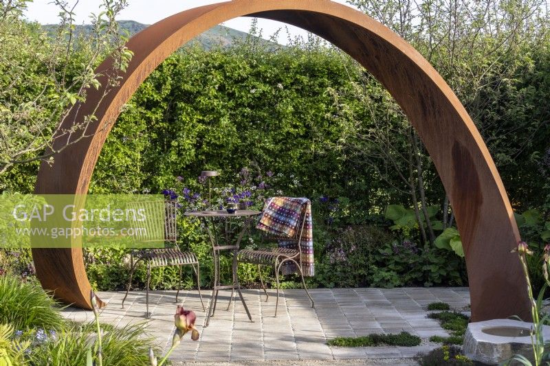 Table et chaises en métal sur pavé sous une arche en acier Corten - 'Ça ne doit pas coûter la Terre' - designer Michael Lote - RHS Malvern Spring Festival 2024 