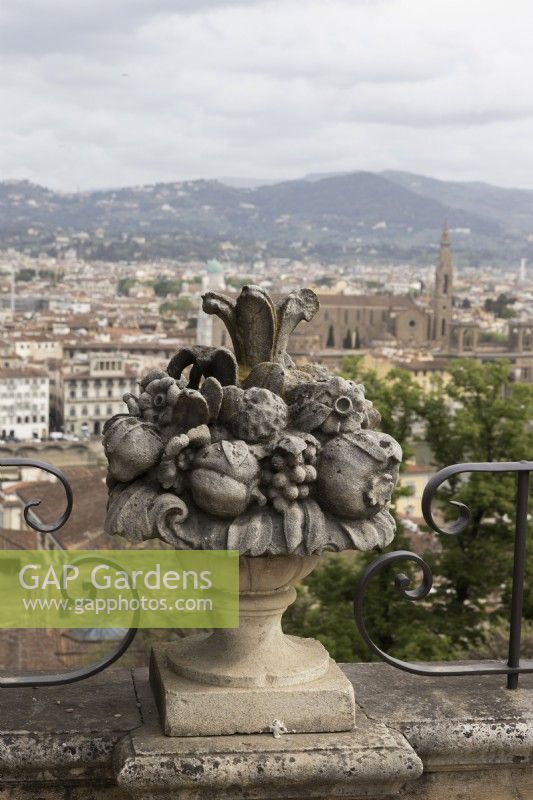 Une sculpture ornée d'une urne pleine de fruits se trouve entre des balustrades métalliques à volutes avec une vue sur les toits de Flornece en arrière-plan. Jardins Bardini. Avril. Printemps. 