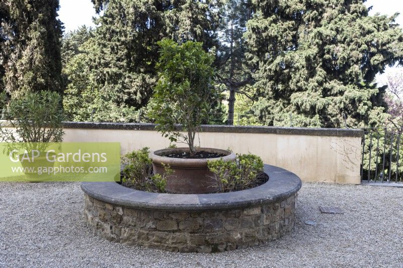Un banc en pierre entoure une grande jardinière en terre cuite sur une terrasse gravillonnée. Jardins Bardini. Florence. Printemps, avril. 
