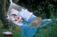 Hamac rayé avec coussins, chapeau et châle et verre de vin blanc avec livre ouvert sur une petite table en bois au Rose Cottage.