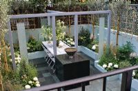 Structure en acier peint gris supportant une table 'flottante' en marbre. Cour - jardin 'divertissant' en blanc, violet et vert. Treillis avec des oliviers formés en forme d'éventail pour l'intimité