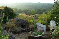 Coin salon avec lavabos alpins, plantes auto-ensemencées, Rosa 'Roserie de L ' Hay' et jardin marécageux avec statue et obélisque. Vue sur la montagne Dinas et le pic Garn Fawr au loin