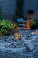 Jardin de nuit avec une rivière de gravier réfléchissante qui court autour de galets, table et chaise en métal en arrière-plan