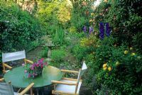 Petit jardin de style cottage de ville avec table et chaises de direction sur terrasse en brique. Delphinium, Campanula, Alchemilla mollis et Roses en parterre de fleurs. Impatiens en pot sur table - Londres
