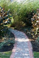 Chemin avec une conception de marguerite en galets et pierres sertis de béton coloré menant à un tunnel de saule avec Corylus avelana 'Purpurea' et des herbes de chaque côté