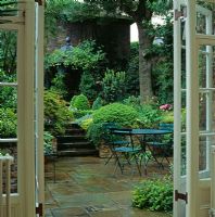 Vue à travers les portes-fenêtres sur la cour pavée, la salle à manger et les marches flanquées d'un feuillage vert luxuriant - Conçu par Jill Billington
