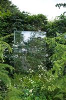 Panneau de verre soutenu par une haie utilisé comme point focal à la fin du chemin avec une plantation de feuillage vert. Design - Amanda Patton pour APL et SGD Le jardin du voyageur - avec Bradstone - RHS Hampton Court Palace Flower Show - Médaille en vermeil