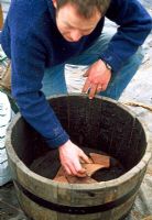 Homme mettant des éclats de terre cuite cassés au fond du tonneau en bois pour faciliter le drainage, avant de le planter