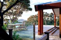 Grande terrasse en bois avec garde-corps en verre maximisant la vue mer. Arbres de Pohutukawa et environs de Manuka