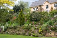 Jardin avant de maison de campagne avec parterres en terrasses abrités et murs en pierre, richement plantés d'alpins, de vivaces, de conifères et d'arbustes - Bank House, NGS garden, Cheshire