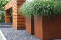 Jardin moderne avec de grandes boîtes rouillées sur un patio surélevé planté d'herbes ornementales. Arche en bois artistique sur l'étang.