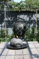 Jardin de méditation urbain. Statue de Bouddha dans un parterre d'Helleborus foetidus. Bougies sur la table au premier plan. Deux arbres Pyrus communis près du mur gris.