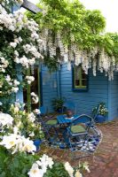 Terrasse carrelée en mosaïque dans un petit jardin avec escalade de roses et de glycines sur pergola. Maison et mobilier peints en bleu. No 11, Christchurch, Nouvelle-Zélande