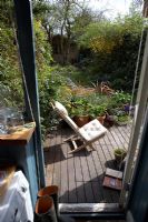 Vue sur le petit jardin de la ville à travers la porte de la cuisine ouverte montrant une terrasse en bois avec une chaise au soleil, des bottes, un livre, des plantes en pots et des plantes plug, avec une vue sur le jardin jusqu'à la maison d'été et les pruniers matures.