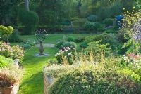 Le jardin supérieur a un cadran solaire en son centre et des parterres de fleurs en forme remplis d'herbes, de plantes vivaces herbacées dont Peonia et d'arbustes intéressants avec étang de moulin vu au-delà. Mill House, Netherbury, Dorset, Royaume-Uni