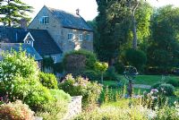 Le jardin supérieur avec cadran solaire et parterres de fleurs en forme d'herbes, de plantes vivaces herbacées, y compris des pivoines et des arbustes intéressants avec étang de moulin vu au-delà - Mill House, Netherbury, Dorset