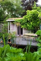 Petit jardin urbain avec terrasse en bois à côté de la maison d'été rose avec des glycines. Étang avec Gunnera qui grandit autour d'elle. Agave en pot.