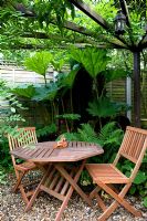 Patio en gravier dans un petit jardin urbain avec table et chaises pliantes en bois sous pergola. Gunnera et Ferns derrière.