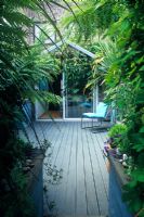 Petit jardin de cour de ville avec terrasse en bois, plantation de style tropical, chaise contemporaine devant véranda. Alistair Davidson, Worcester, Royaume-Uni