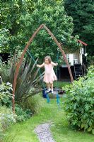 Fille jouant dans le jardin de la ville sur une balançoire suspendue à une arche métallique. Phormium sur le côté gauche et cabane en bois derrière. Jardin Yulia Badian, Londres, Royaume-Uni