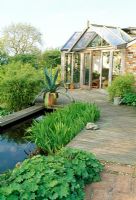 Terrasse en bois et brique, étang et véranda. Agave en pot. Fin du printemps, Fovant Hut Garden, Wilts