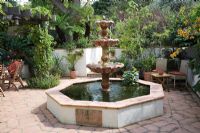 Fontaine à gradins en marbre italien en piscine surélevée octogonale sur carreaux de terre cuite. Vigne sur pergola en chêne et chaises en osier dans un petit jardin urbain