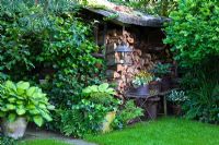 Pile de bois dans un abri de jardin avec lanterne suspendue, table pliante en métal avec pot de fleur, boîte et Hosta en pots en terre cuite et en osier