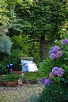 Chaise en fer en bois avec coussins bleus sur une aire de repos pavée ronde avec des pots en terre cuite et un ornement de lièvre entouré par Acer platanoides 'Globosum', Buxus et Hydrangea macrophylla