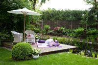 Mobilier de jardin en osier blanc, une couverture avec des coussins et un parasol sur une terrasse en bois à côté d'un étang de jardin. Treillis de saule et haie en arrière-plan. Les plantes sont coupées Buxus, fougère et hortensia