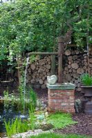 Un puits d'eau à côté de l'étang, avec deux escargots en pierre sculptée, des urnes en métal et un tas de bois en arrière-plan. Les plantes comprennent Lysimachia nummularia, Salix et Typha