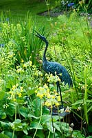 Statue de héron à côté de l'étang entouré de Primula grandiflora, Iris, Alchemilla mollis et fougères - Mindrum, nr Cornhill on Tweeds, Northumberland, UK