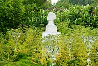 Tête de pierre de Portland sculptée par l'artiste local Tom Clark, parmi une masse d'Eryngium giganteum, fougère Ligusticum lucidum sur fond de Phytolacca americana blanc - Yews Farm, Martock, Somerset, UK