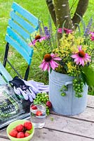 Sur une table en bois un vase avec coneflower, Ladys Mantle, Veronica et raisins. Également sur la table, un livre de jardinage, des gants de jardinage, une fourchette à main, un bol de fraises et un verre d'eau. En arrière-plan une chaise de jardin turquoise colorée.