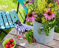 Sur une table en bois un vase avec coneflower, Ladys Mantle et raisins. Également sur la table, des gants de jardinage, une fourchette à main, un bol de fraises et un verre d'eau. En arrière-plan une chaise de jardin turquoise colorée.