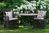Deux fauteuils en osier avec coussins patchwork brodés et une table de bistrot avec bougeoirs sur fond de fleurs blanches Hydrangea arborescens 'Annabelle'
