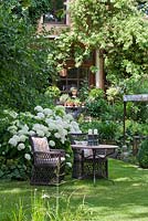 Deux fauteuils en osier avec coussins patchwork brodés et une table de bistrot avec bougeoirs sur fond d'Hortensia arborescens à fleurs blanches 'Annabell ''.
