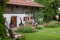 Chaises de terrasse en bois sur une pelouse devant une maison bavaroise de style traditionnel avec volets et rosace grimpante. Les plantes sont Rosa 'Grusse an Bayern', Rosa 'New Dawn', Centranthus ruber 'Albus', Centranthus ruber 'Coccineus' et Petunia