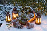 Nature morte sur le thème de Noël dans la neige avec un trug en bois contenant des pots de fleurs et des pommes de pin, des lanternes et du gui