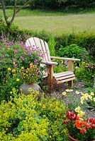 Chaise de jardin Adirondack entourée de Geranium psilostemon et Alchemilla mollis. Pots avec Pelargoniums, Altos, Nemesias et Marguerites. Haie avec prairie en arrière-plan