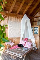 Sous un porche en bois, un chat noir est assis sur un transat avec des coussins et une moustiquaire