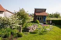 Jardin de campagne moderne avec cabanon en bois, meubles de jardin anciens sur une pelouse, un mur en pierre des champs, tas de pierre et parterres de fleurs mixtes, Viburnum plicatum, haie de cerisier