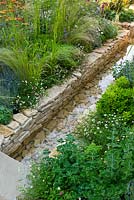 Un canal d'eau entouré de plantations de vivaces, d'herbes et d'arbustes dans le jardin 'Four Corners' conçu par Peter Reader au RHS Hampton Court Flower Show.